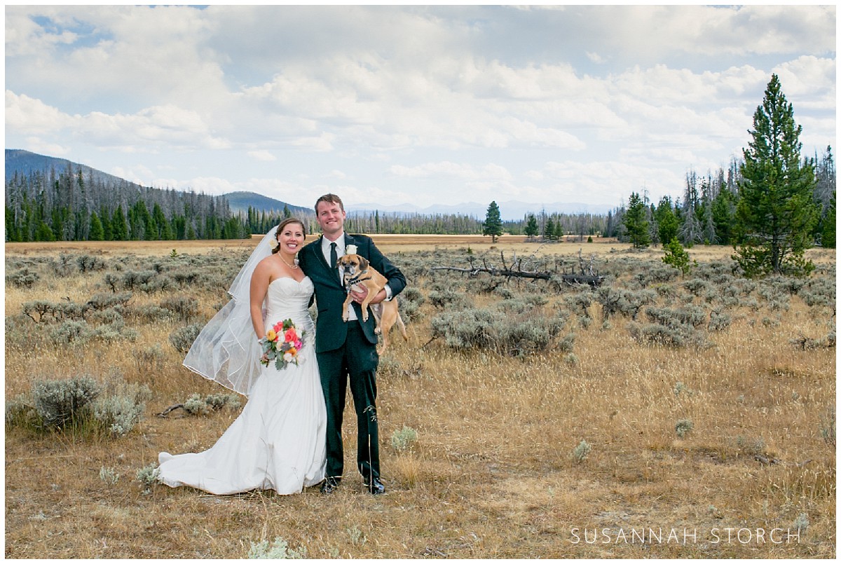 a bride and groom stand in a western landscape while holding their small dog