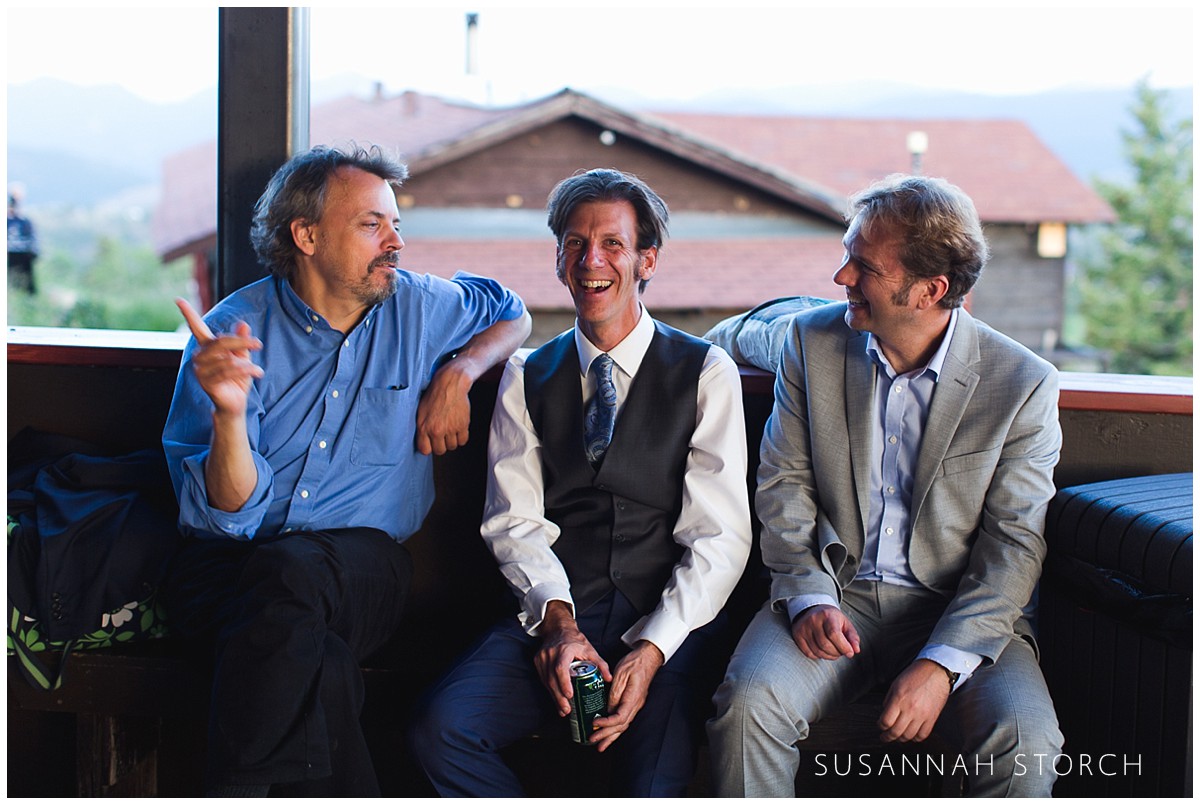 Three men (one a groom) smile and chat at sunset on a deck.