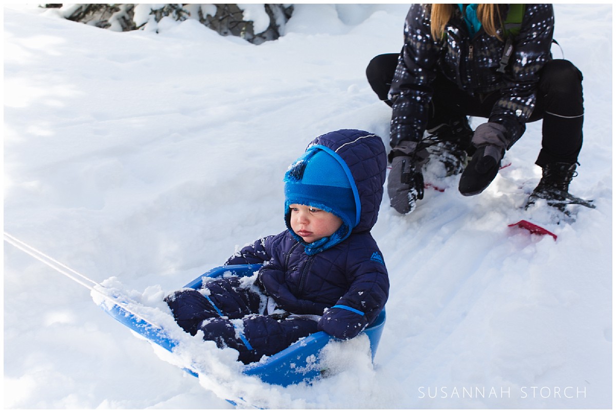 a mom stands behind her young son as he sleds down a gentle hill