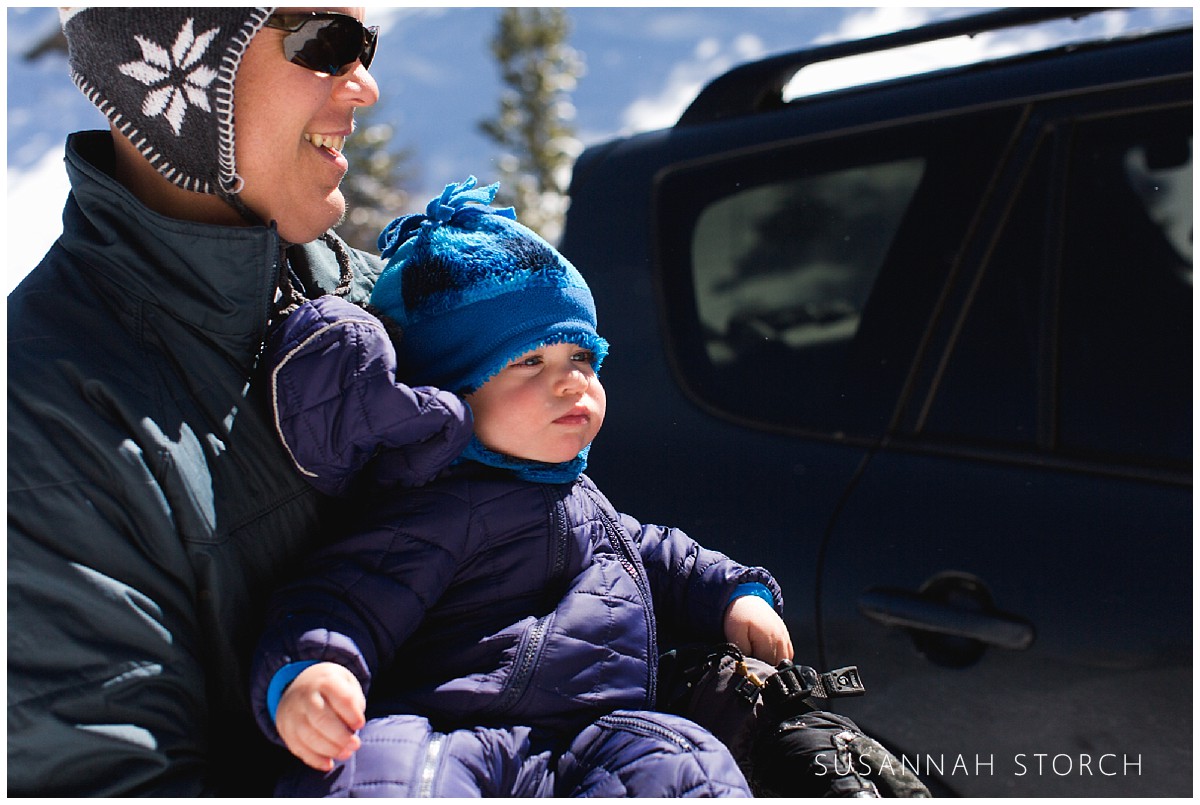 a toddler in a snowsuit sits on dad's lap