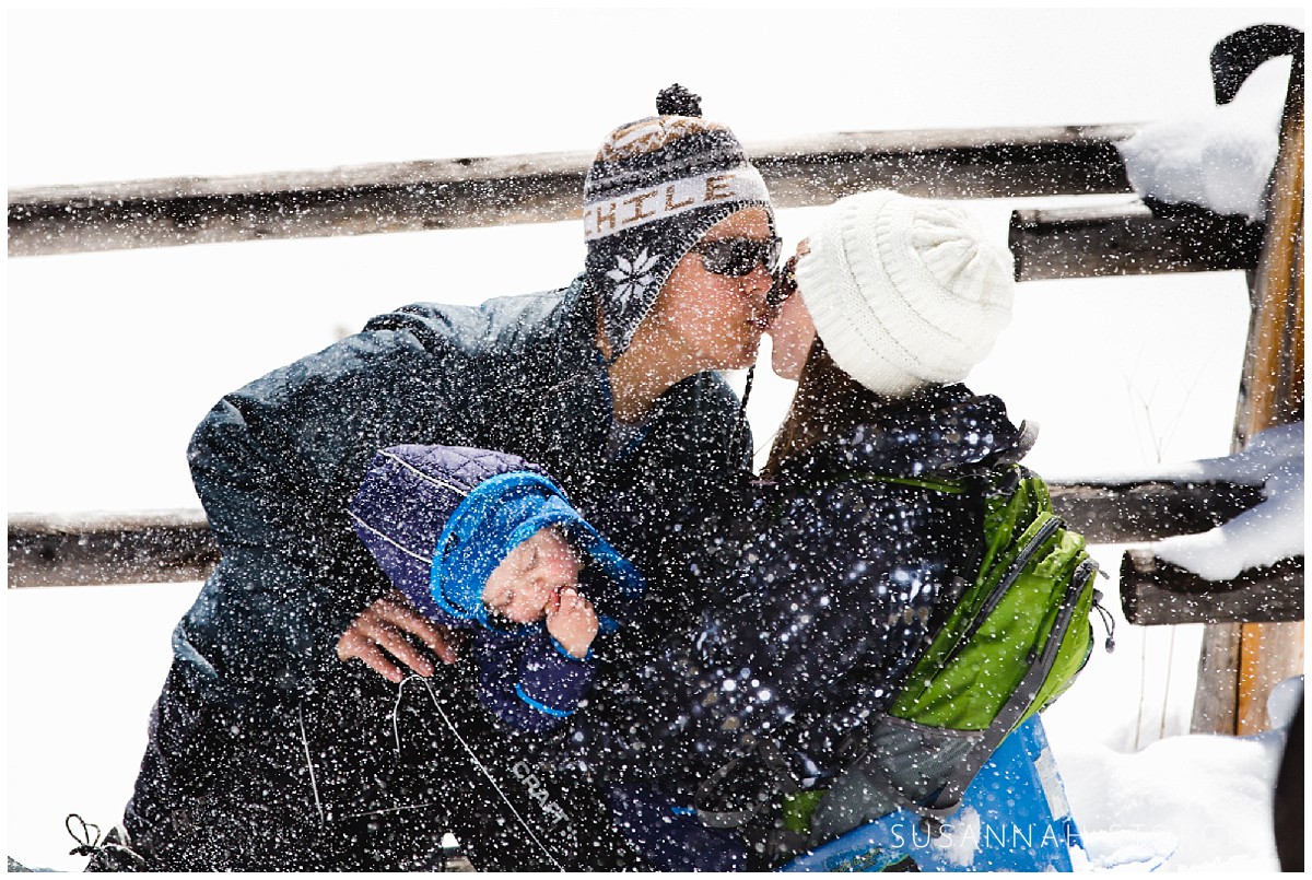 a husband and wife kiss while holding their young son in the snow