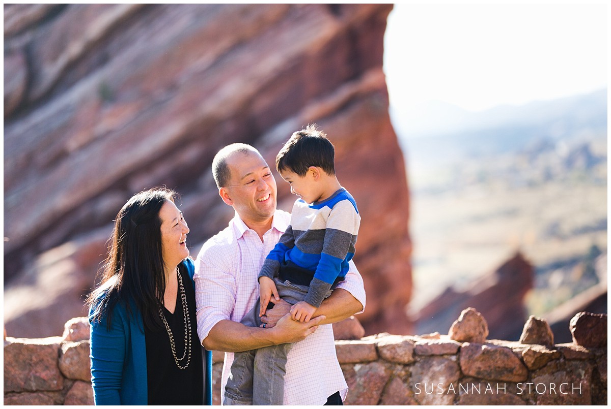 A dad holds his son as mom watches in this photograph in front of a red rock wall