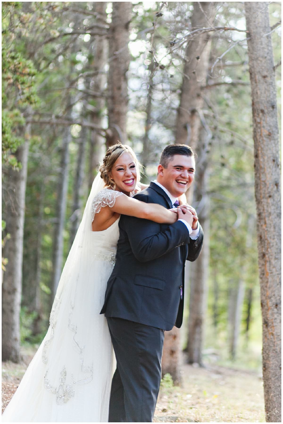 a bride bear hugs her new husband in front of pine trees