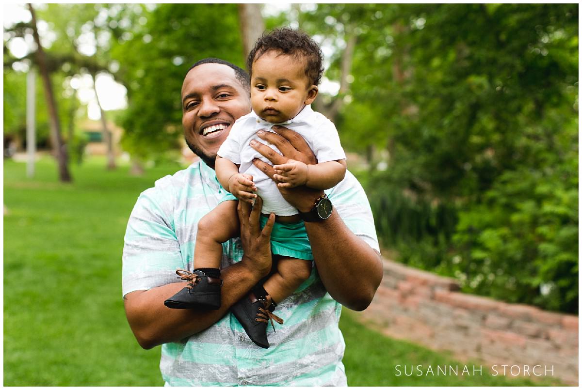 smiling father hold his sweet baby son in front of green bushes and trees