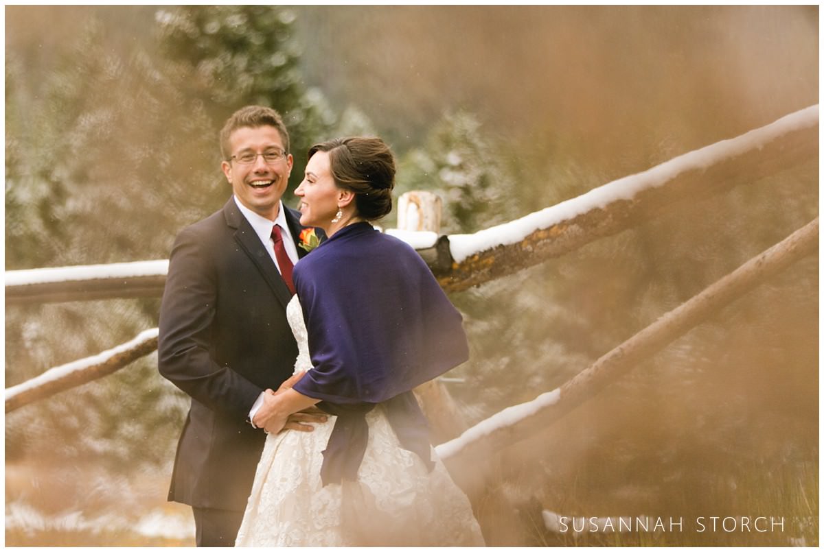 a bride and groom stand by a wooden fence on a snowy fall day