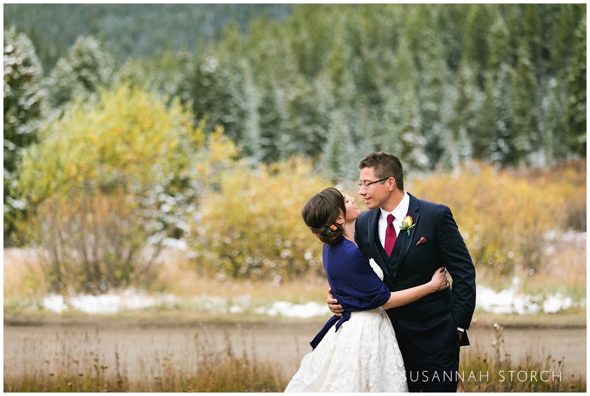 a couple look at each other while standing in front of snowy pine trees