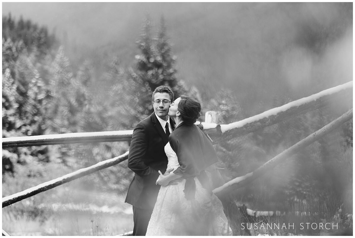 black and white image of a bride kissing her groom among pine trees