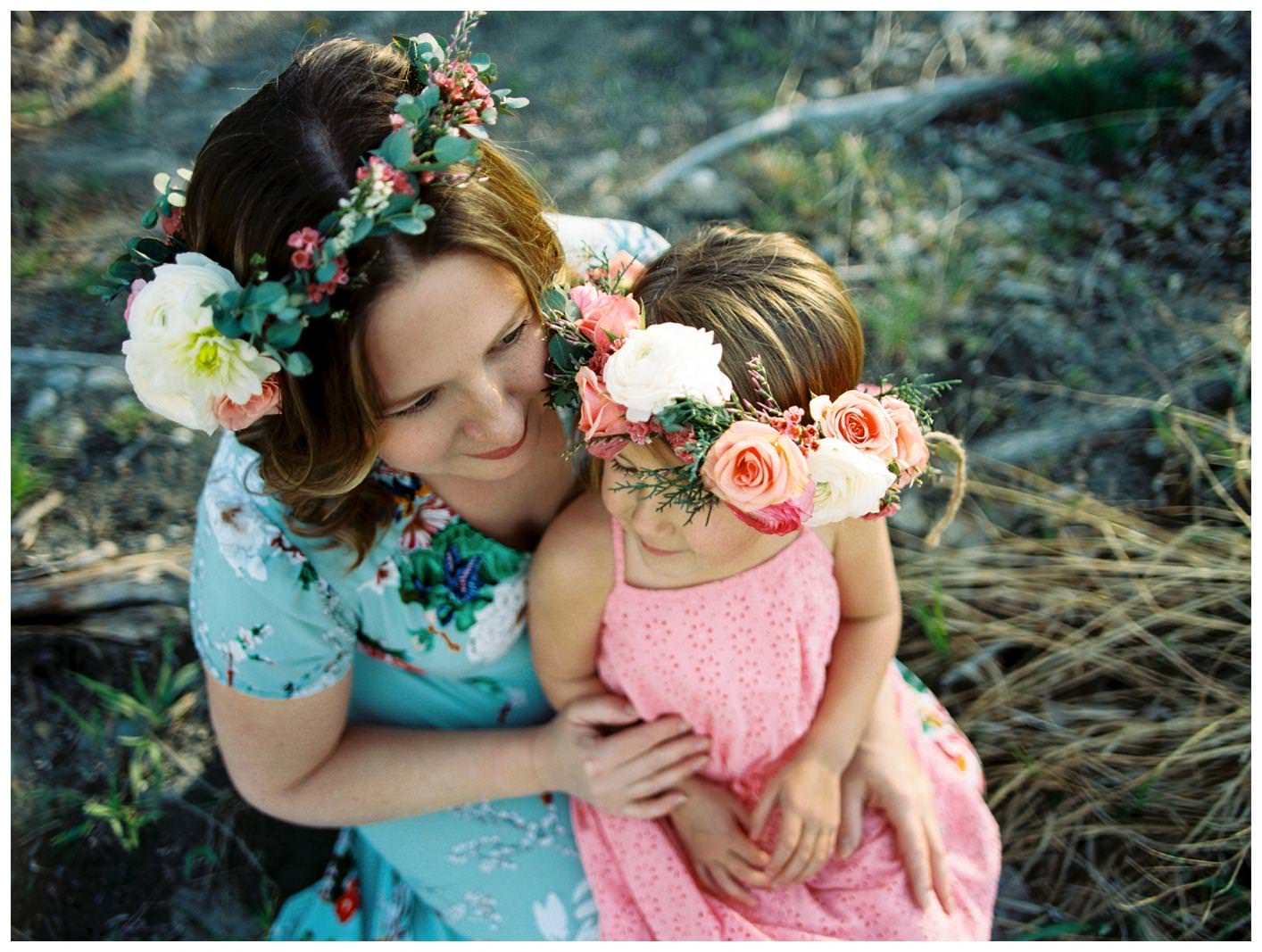 A daughter sits on her mama's lap and both wear floral crowns