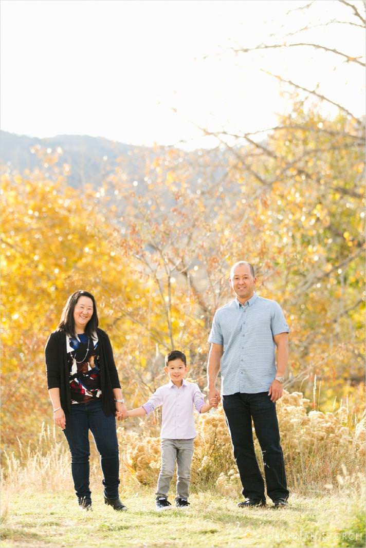 mom, son, and dad hold hands in front of fall trees and mountains in golden, colorado during a family photo session