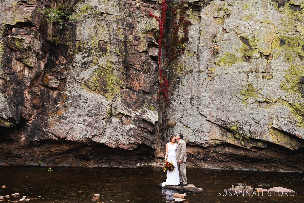 a bride and groom stand on a rock in a river in front of a big rock wall