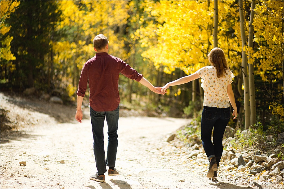 couple hold hands and walk down a dirt road surrounded by aspen trees
