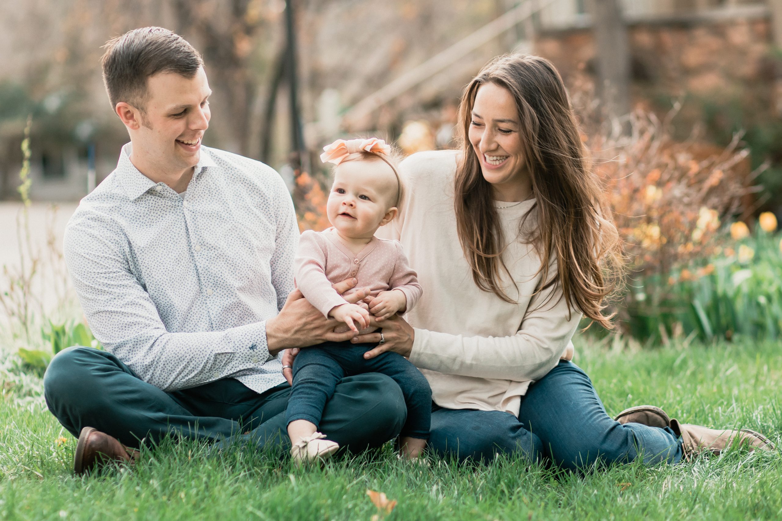 mom and dad smile at baby girl while all three sit on green grass