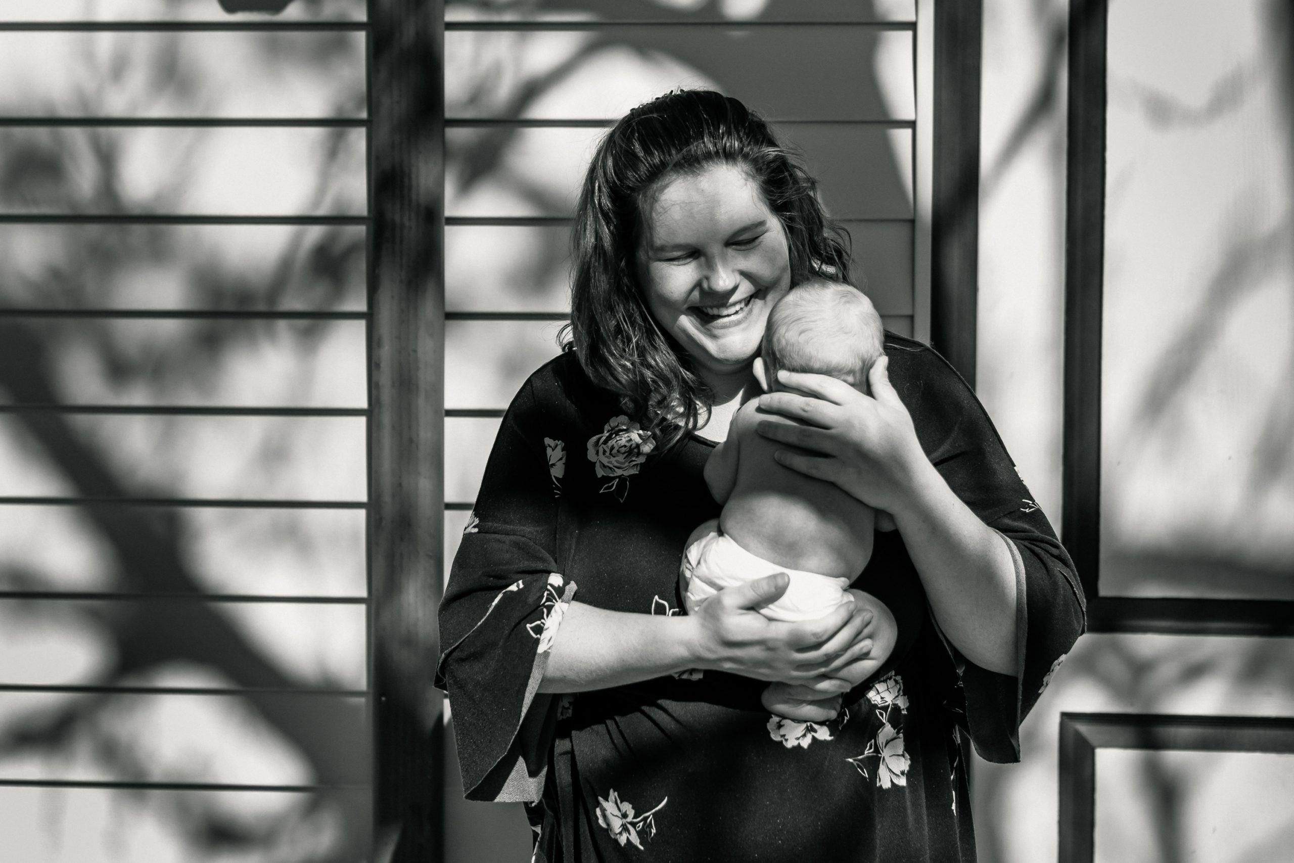 image from blog post on when to schedule a newborn photography session. photo shows a mom holding her baby boy in front a wall with a tree shadow across face.