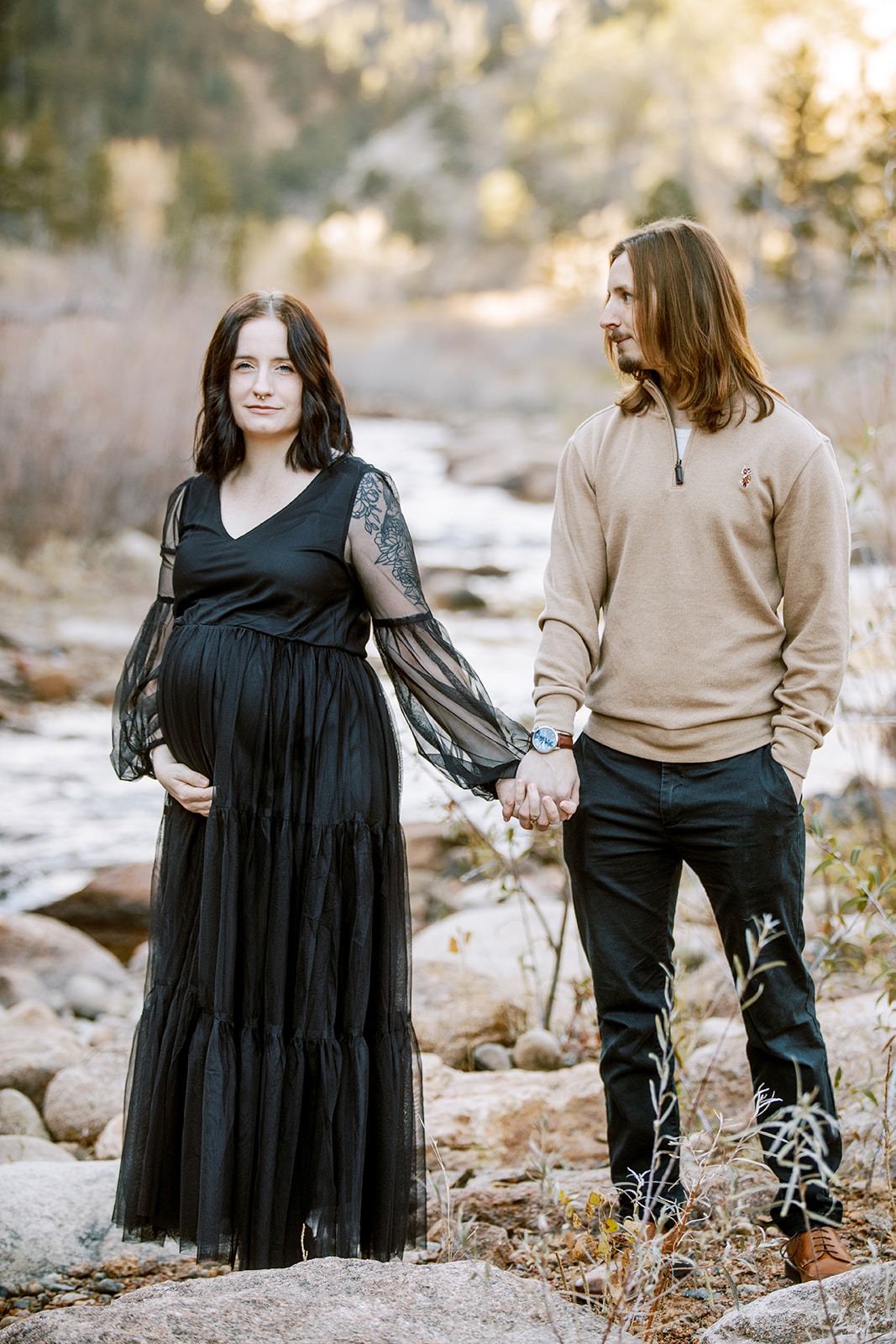a husband looks at his pregnant wife who wears a black long dress and holds belly bump while both stand in front of rocky river
