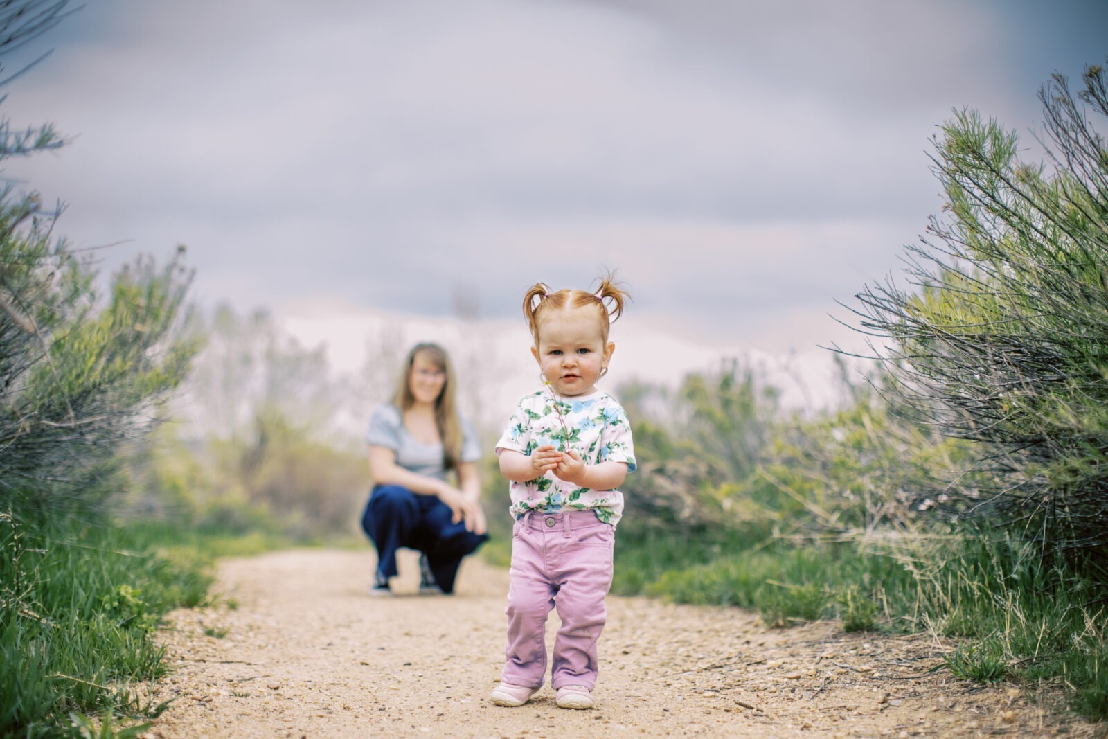 A toddler girl with pigtails stands on a dirt path in front of her mother. Both are surrounded by sage brush.