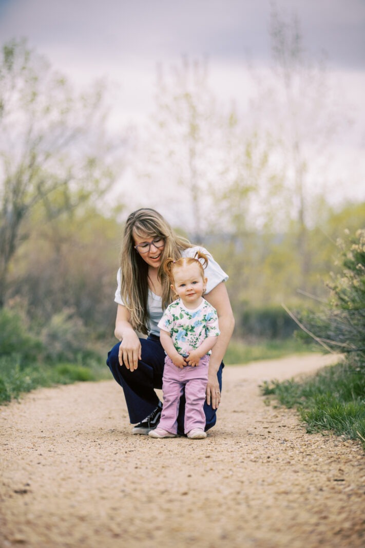 a mom kneels behind her toddler girl who stands on a dirt path