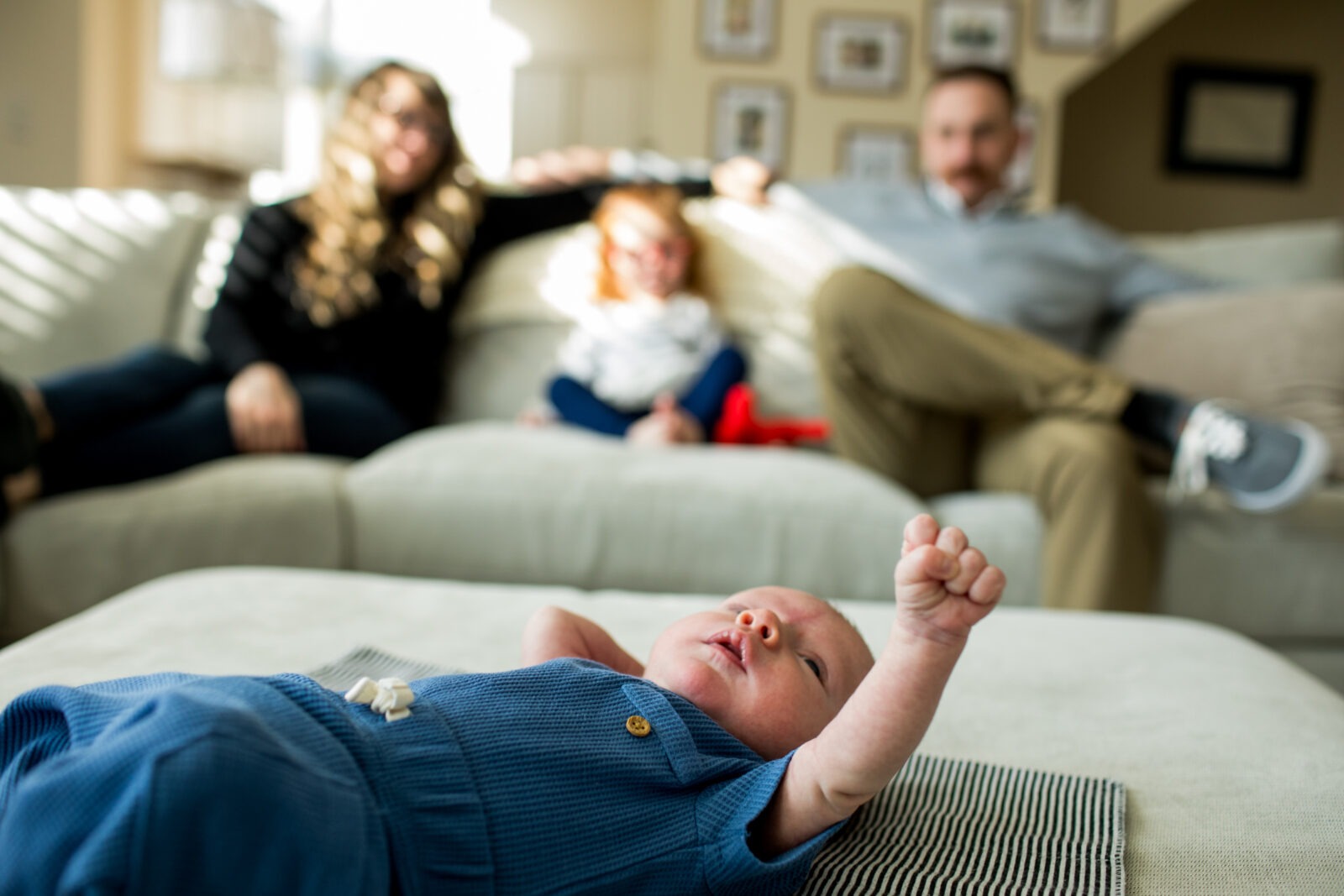 newborn baby lies on a pouf and his parents and sister sit on couch behind him