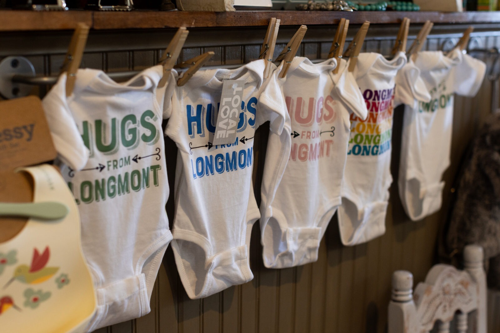 Hugs from Longmont” baby onesie hanging in front of a brick wall at Adornables, a woman-owned baby boutique in downtown Longmont, Colorado, featuring locally inspired gifts and baby clothes.