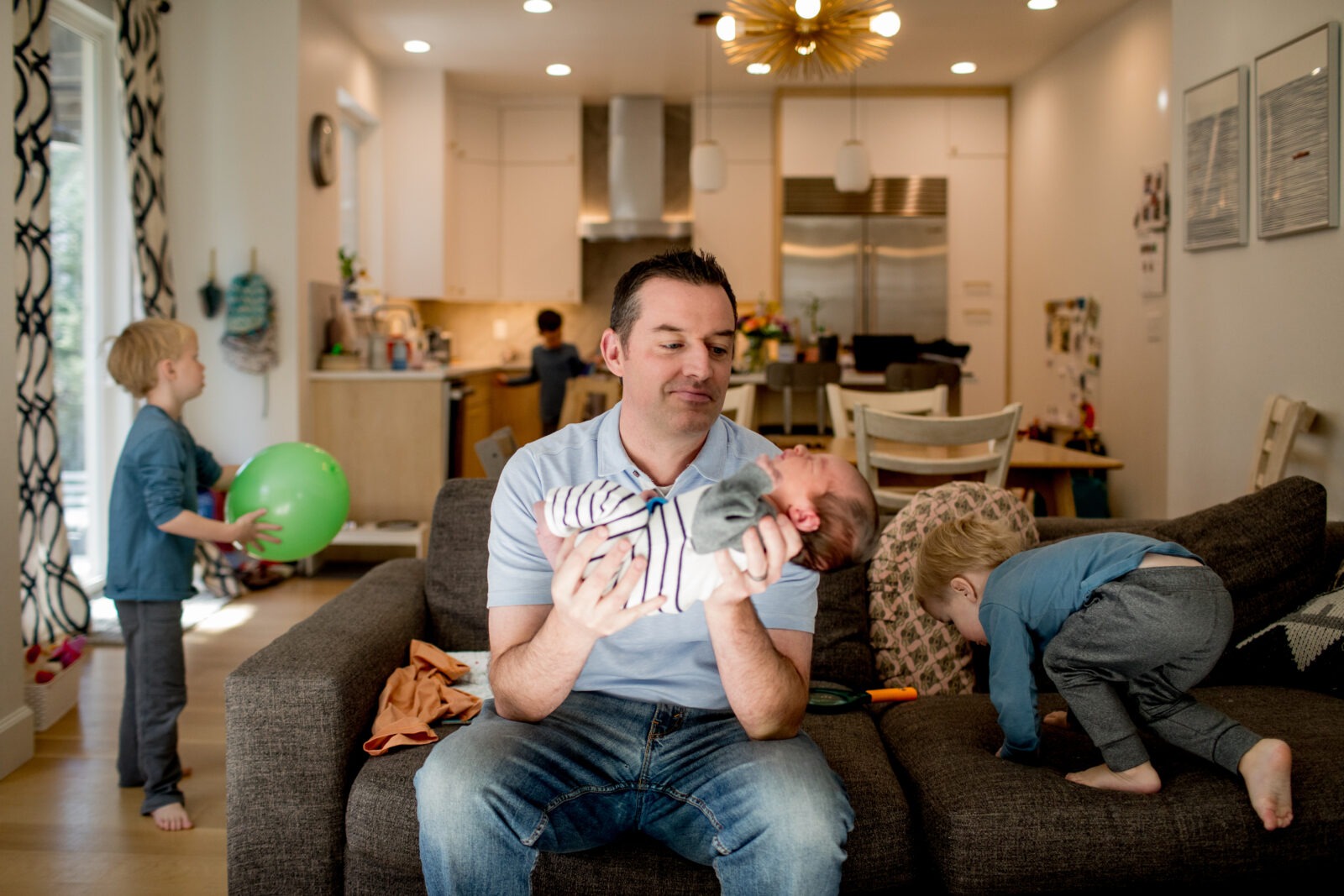 dad holds newborn baby boy in living room while older siblings are active around him